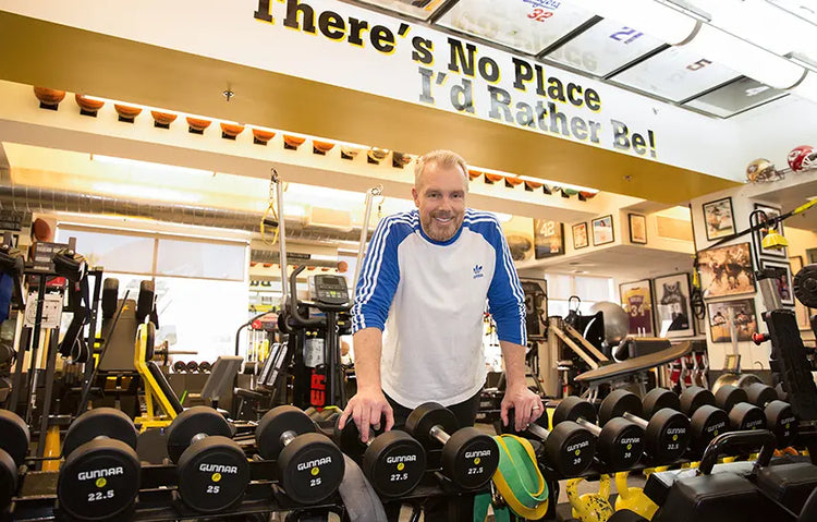 Gunnar Peterson standing among dumbbells with a motivational quote on the wall in a gym.