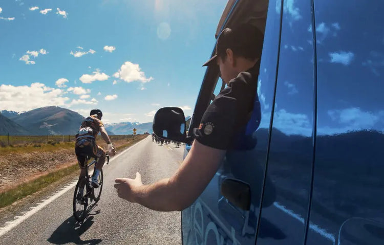NZ cycling project cyclist on a bicycle being waved by a person in a car on a road with mountains in the background